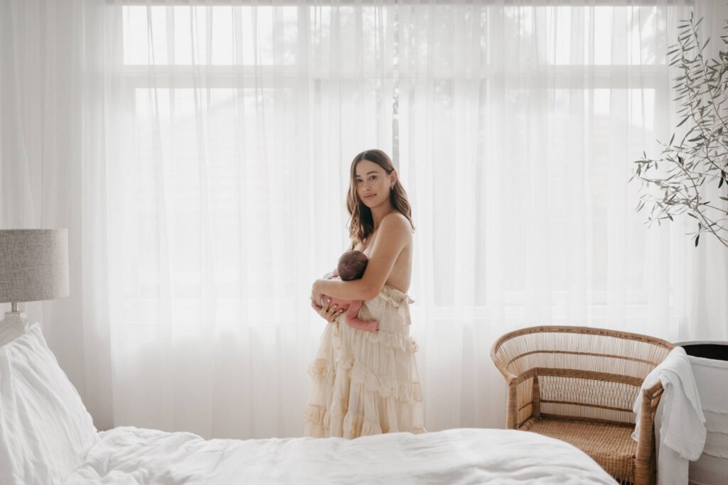 In-home newborn session in Sydney with mother holding her baby in soft window light in a bedroom