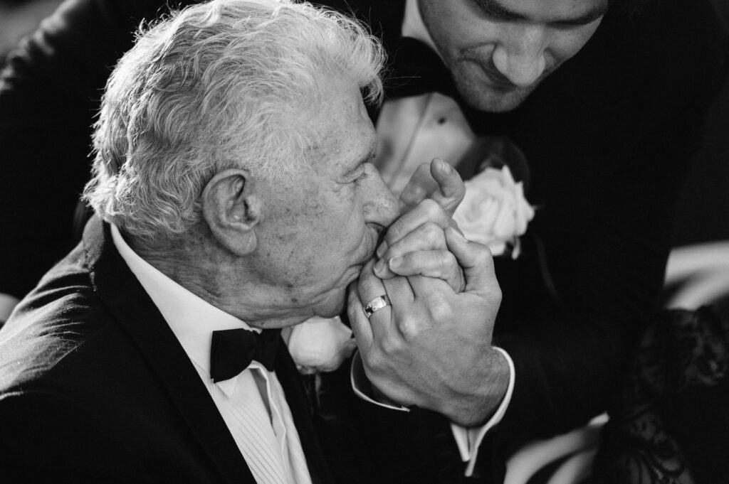 Black and white photograph of an older gentleman tenderly kissing a groom's hand — an emotional wedding moment captured by Tealily Photography Sydney