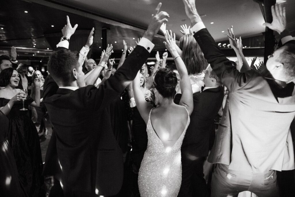 Black and white image of wedding guests celebrating with arms raised on the dance floor — joyful reception moment photographed by Tealily Photography Sydney