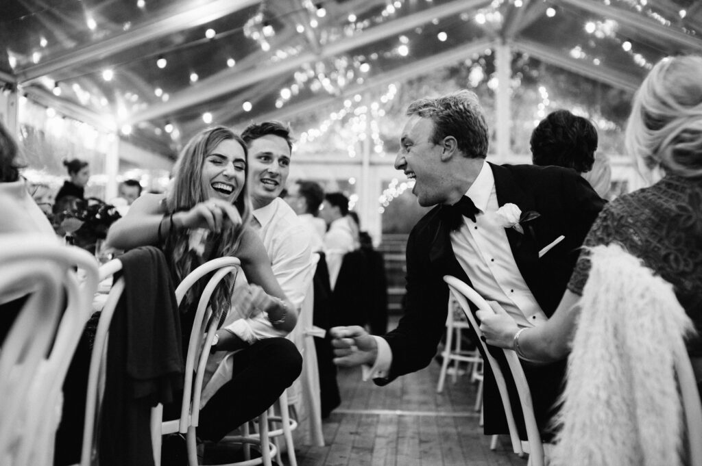 Black and white photograph of guests laughing and dancing under festoon lights at a tented wedding reception — Sydney wedding documentary photography by Tealily