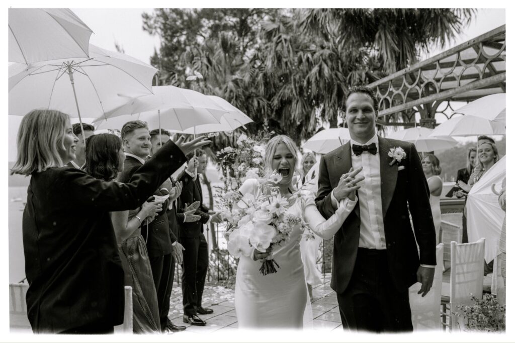 Black and white photograph of a rainy day wedding ceremony — couple walking under umbrellas with guests, joyful candid wedding photography Sydney by Tealily Photography
