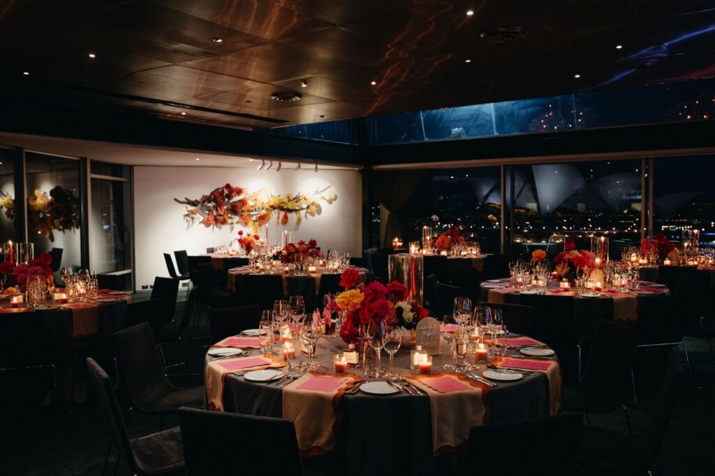 Elegant candlelit wedding reception room at Quay Restaurant Sydney with round tables, red floral arrangements, and a warm, intimate glow — Sydney harbour wedding photography by Tealily Photography