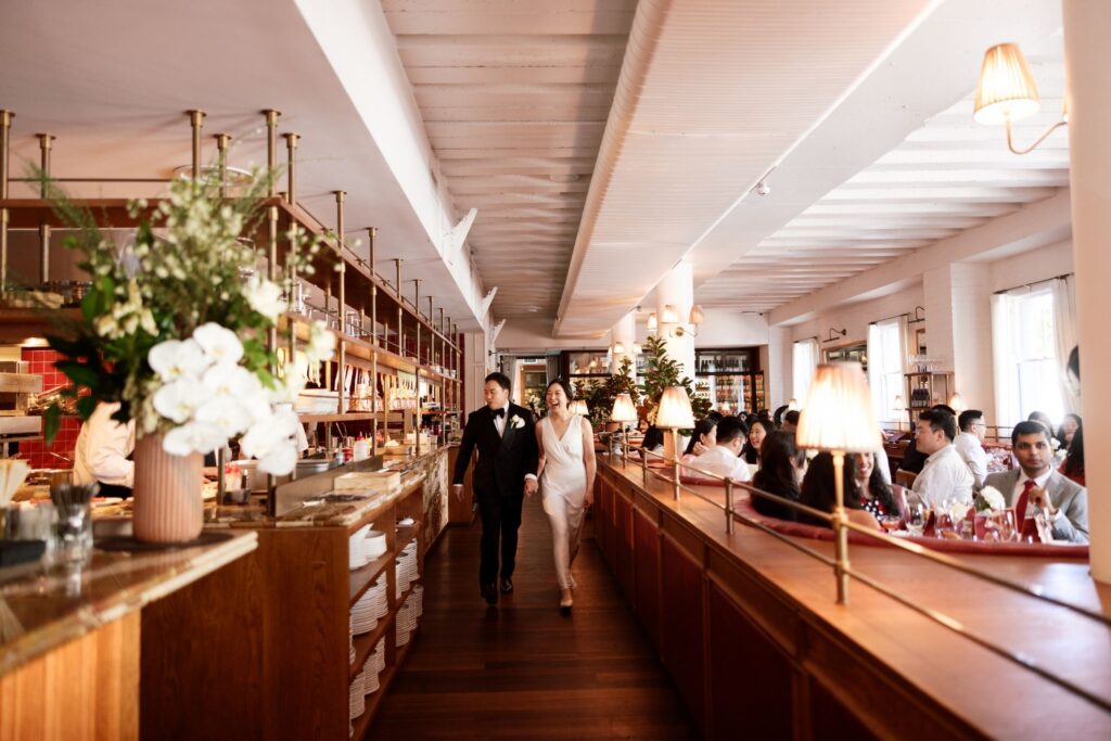 Long dining room at Armorico Surry Hills with guests seated along communal tables, warm pendant lighting, and a couple walking toward the camera — documentary wedding photography by Tealily Photography