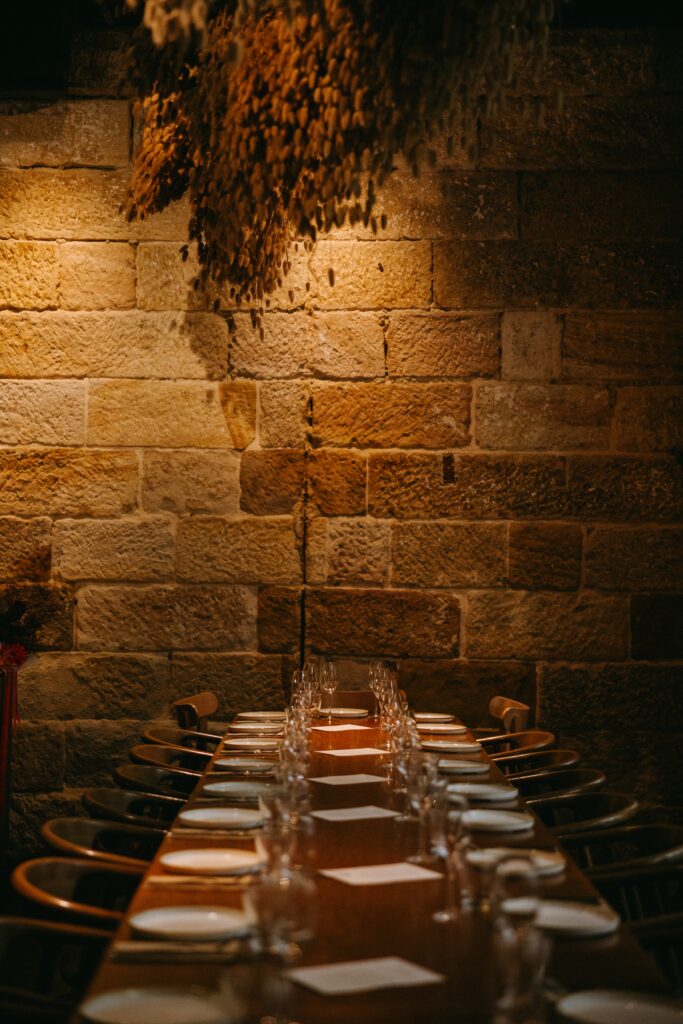 Warm candlelit interior of Grana restaurant showing long tables set for dining beneath dried botanicals and exposed brick walls — intimate Sydney wedding venue near Circular Quay