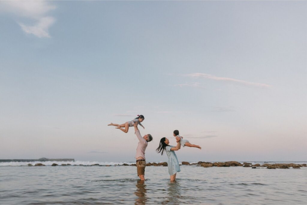 A landscape image of a mum and dad standing against a sunset throwing their daughters into the air as the water laps at their feet during a relaxed family photoshoot