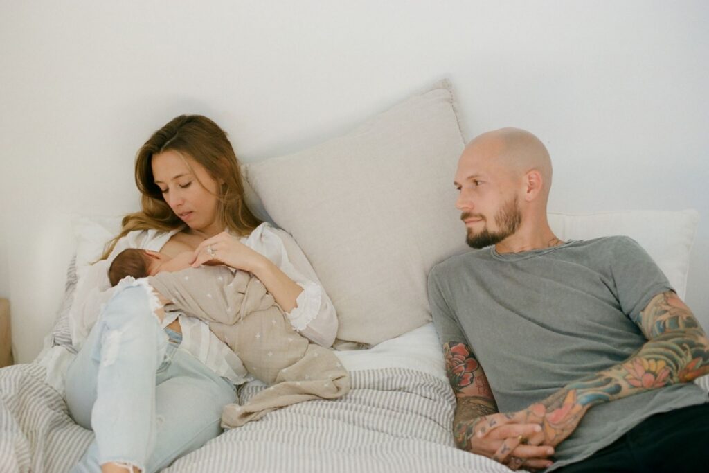 A soft documentary image of new parents at home in bed with their newborn as she breastfed by her mother