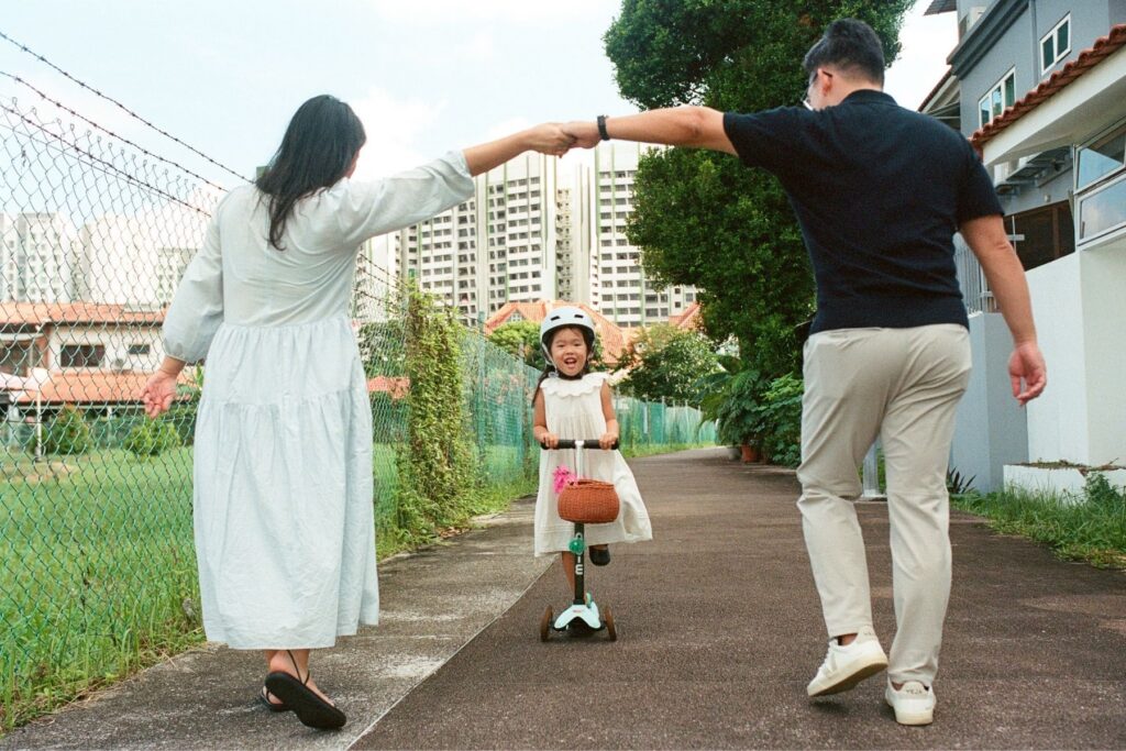 A young girl in a helmet on a scooter going through her parents arms held together above her head during a documentary family photoshoot