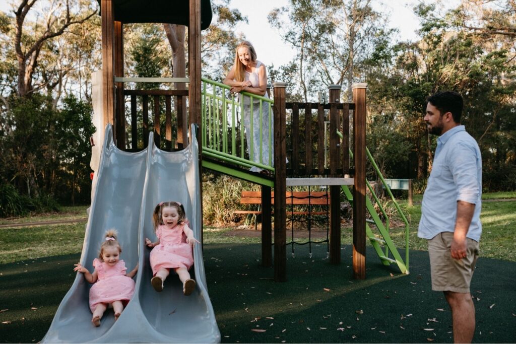 A family relaxing at their local park during a documentary family session, two girls in pink happily going down a slide while their parents watch on